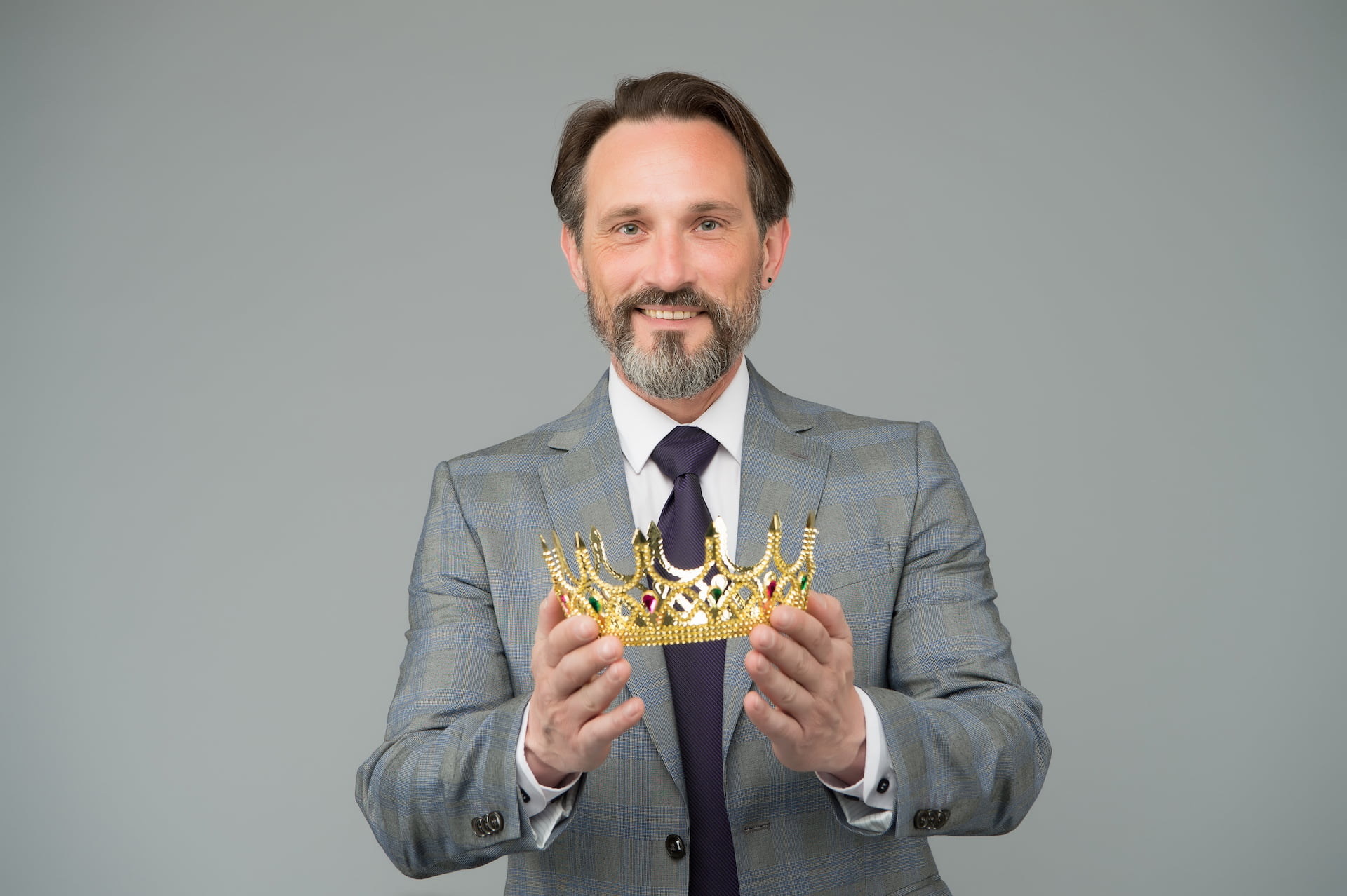 Lachender Mann mit einer Krone in der Hand vor grauem Hintergrund.